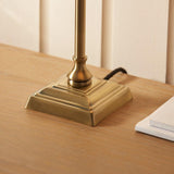 Close-up of the Endon Caldmont table lamp's antique brass square stepped base and slender column on a light wood surface beside white notebooks.