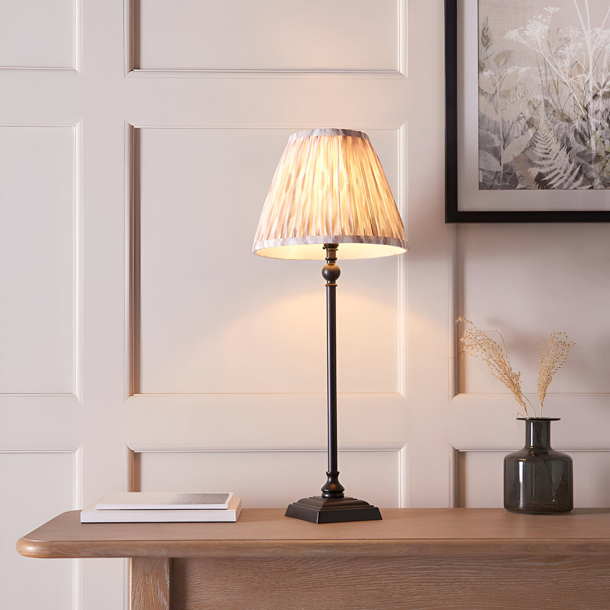 Caldmere Ikat Table Lamp in cream, with a pleated fabric shade on a slim matt black metal candlestick base, shown on a console table in a living room setting.