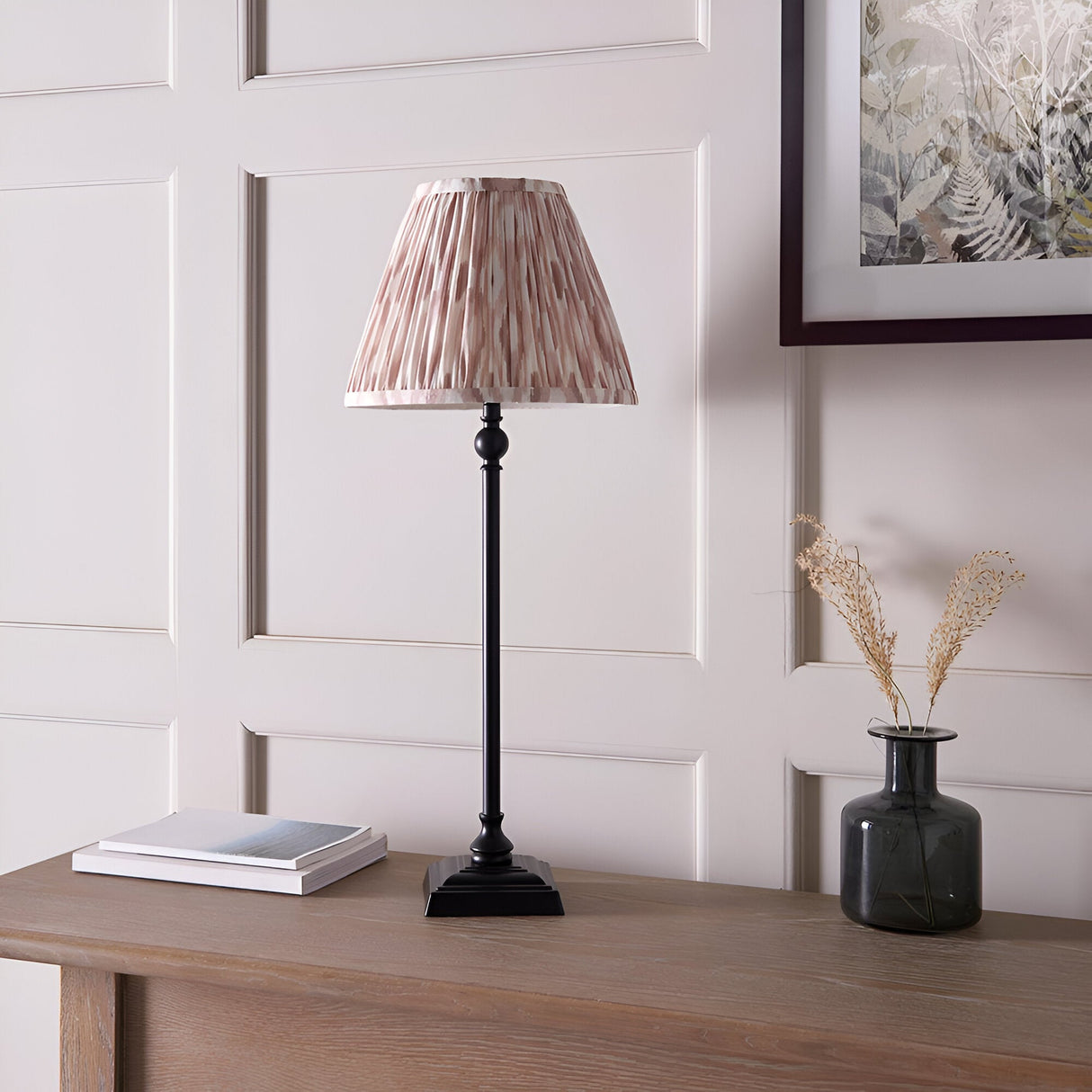 Caldmere Ikat Table Lamp with slim matt black candlestick base and pleated cream-toned patterned shade, shown on a wooden console beside a vase and books.