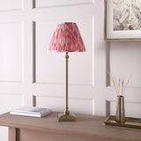 Caldmere Ikat Table Lamp in coral, with a pleated fabric shade on an antique brass candlestick-style base, styled on a wooden console beside a mirror and vase.