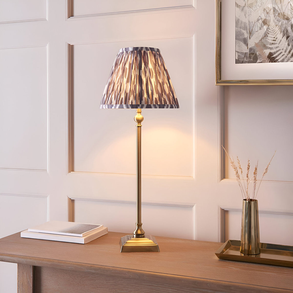 Endon Caldmere table lamp with slim antique brass candlestick stem and pleated grey Ikat print shade, styled on a wood console beside books and a brass vase.