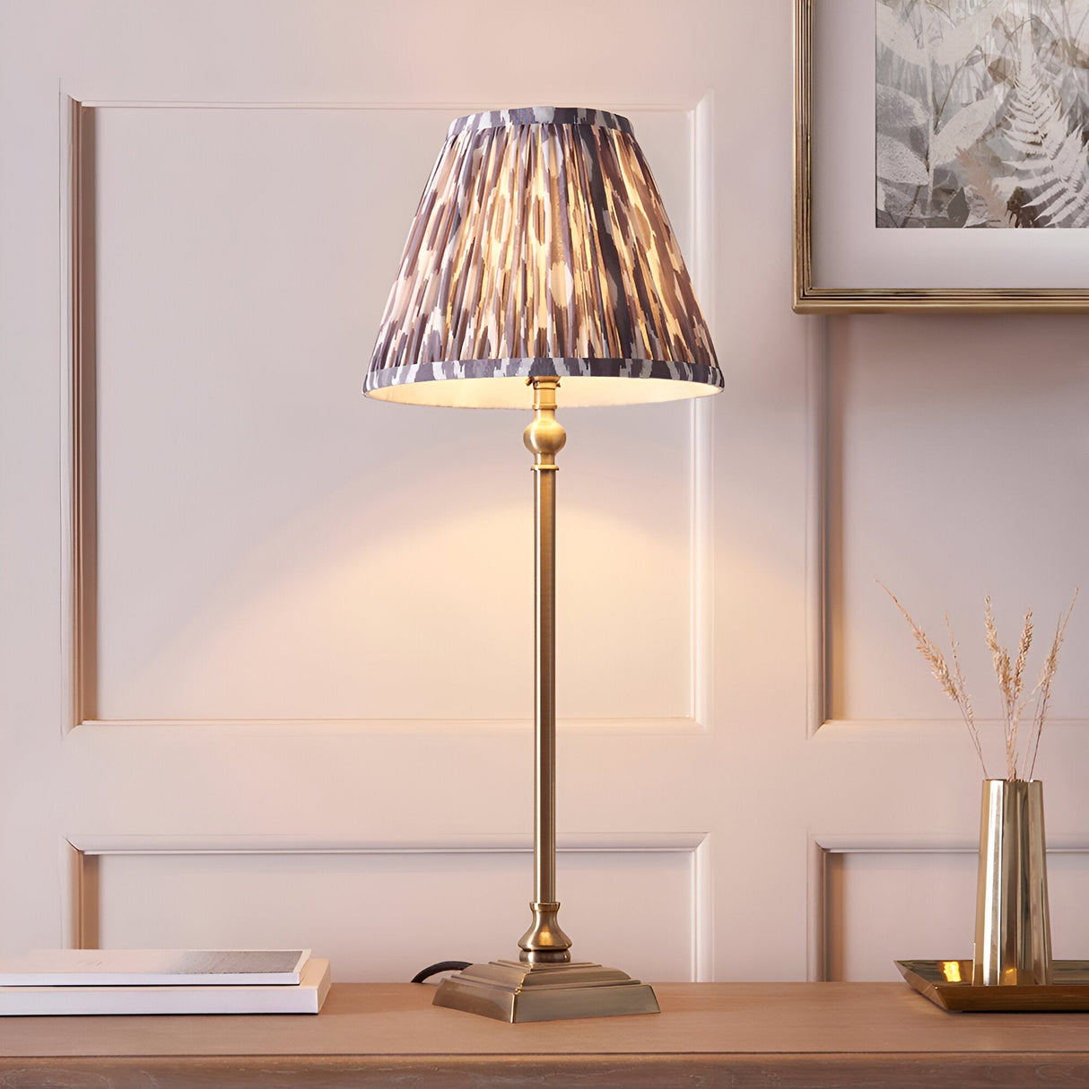 Endon Caldmere table lamp with slim antique brass candlestick stem and pleated grey Ikat print shade on a wooden sideboard against panelled walls.