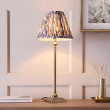 Endon Caldmere table lamp with slim antique brass candlestick stem and pleated grey Ikat print shade on a wooden sideboard against panelled walls.