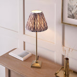 Endon Caldmere table lamp with slim antique brass candlestick base and pleated grey Ikat print shade, on a wood console beside books and a brass vase.