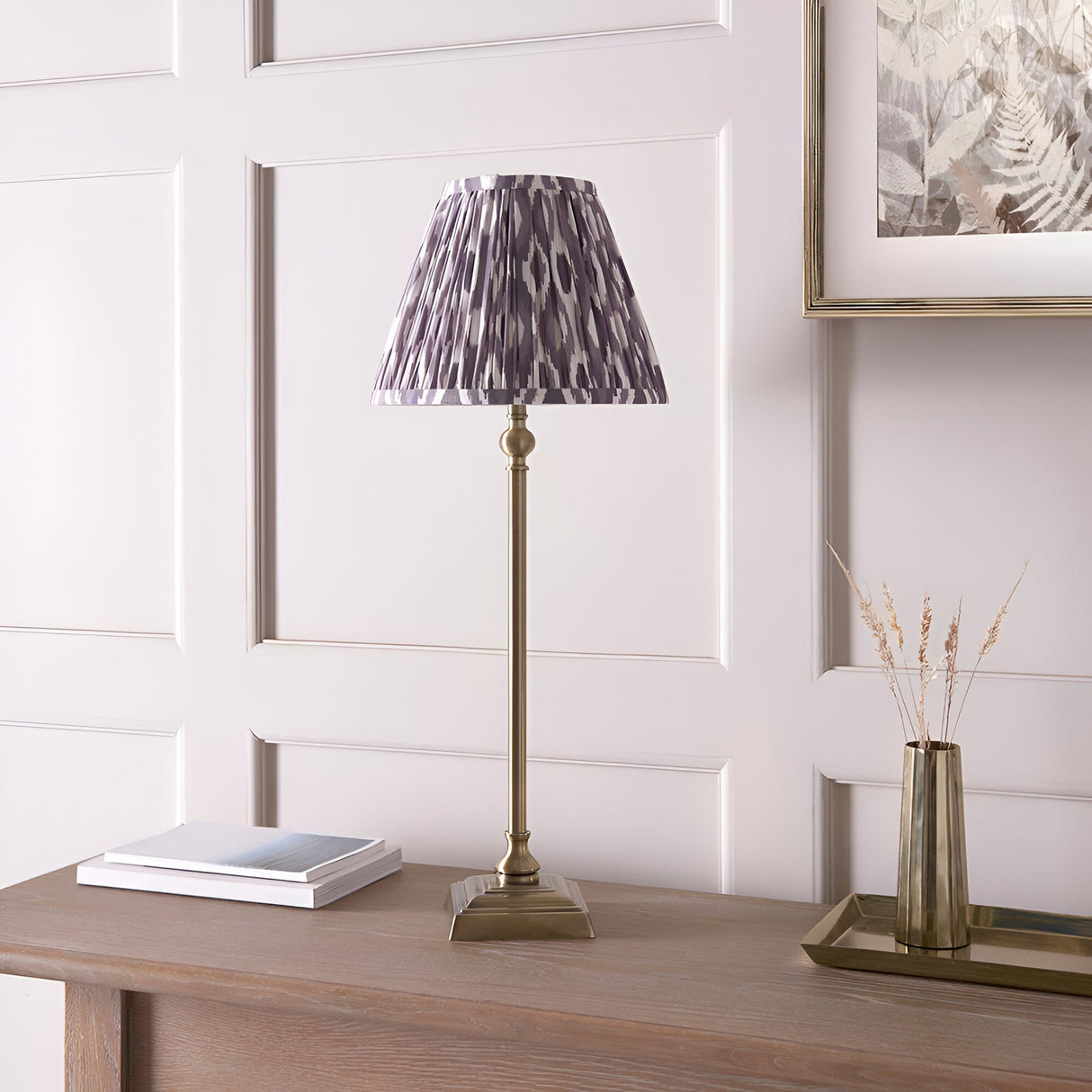 Endon Caldmere table lamp with slim antique brass candlestick stand and pleated grey Ikat shade, styled on a wooden console beside books and a vase.