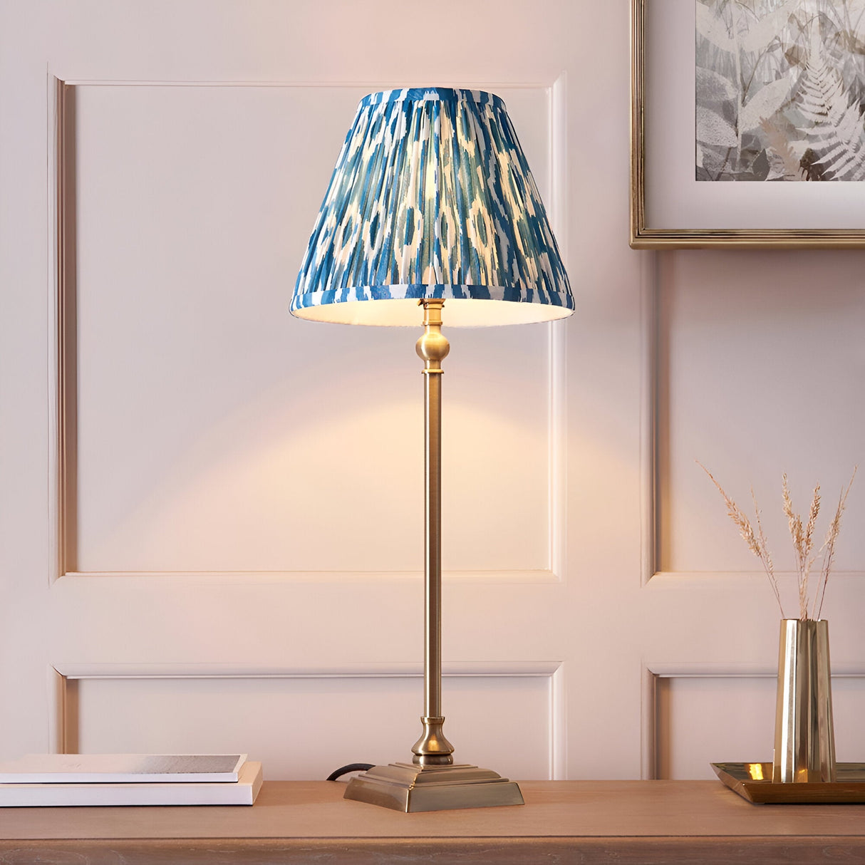 Endon Caldmere table lamp with slim antique brass candlestick stem and pleated blue Ikat print shade, on a wood console beside books and a brass vase.