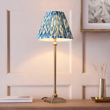 Endon Caldmere table lamp with slim antique brass candlestick stem and pleated blue Ikat print shade, on a wood console beside books and a brass vase.