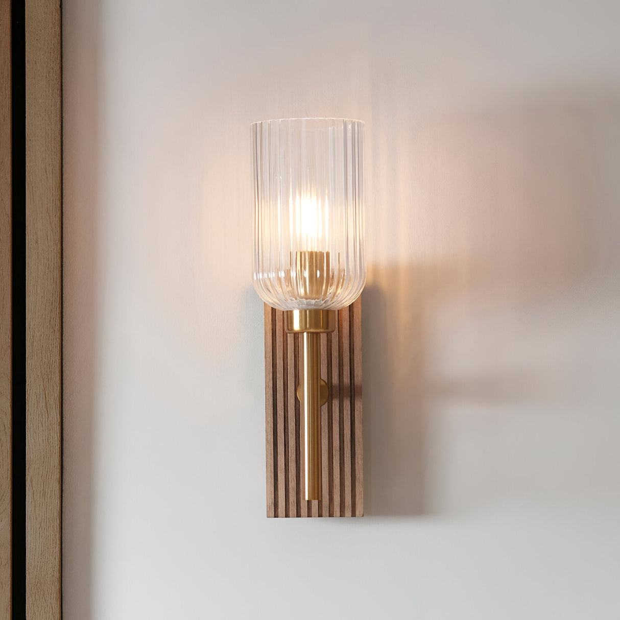 Bramton Wall Light in brass with a ribbed clear glass shade and grooved walnut-stained wood backplate, mounted lit on a plain wall.