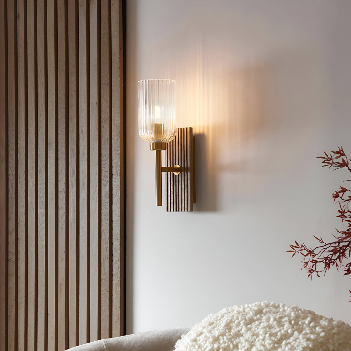Bramton Wall Light in brass with a ribbed glass shade and grooved wood backplate, glowing warmly on a wall beside slatted panelling in a living room.