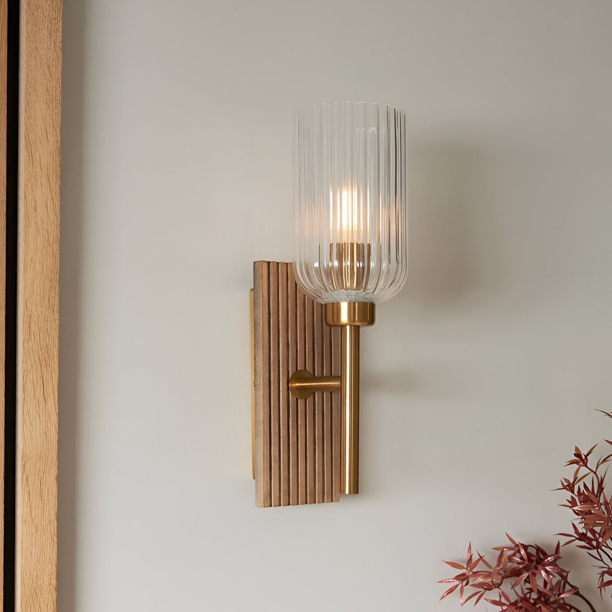 Bramton Wall Light in Brass with grooved wood backplate, brass arm and ribbed glass shade, mounted on a plain light wall.