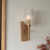 Bramton Wall Light in Brass with grooved wood backplate, brass arm and ribbed glass shade, mounted on a plain light wall.