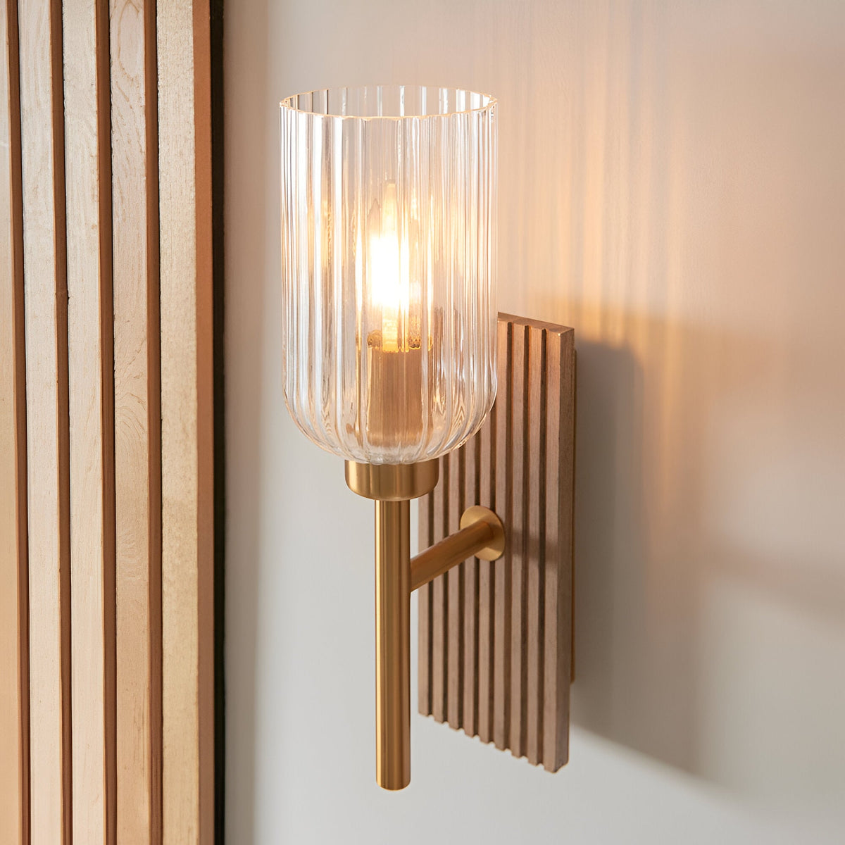 Bramton wall light in brass with a ribbed glass shade, mounted on a grooved wood backplate against a light wall.