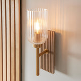 Bramton wall light in brass with a ribbed glass shade, mounted on a grooved wood backplate against a light wall.