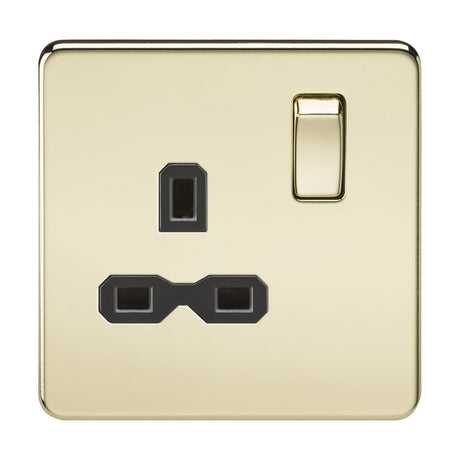 The 13A 1 Gang DP Switched Socket in polished brass features a screwless flat plate with a gold finish and includes a black insert. It has three rectangular pin slots configured in the standard triangular layout for UK sockets, with the switch conveniently placed to the right of the pin slots.