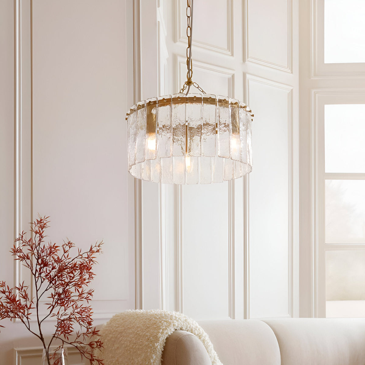 Brockhurst 3 Light Pendant in brass with drum-shaped textured glass panels, hung from a chain in a light-toned living room with a cream sofa nearby.