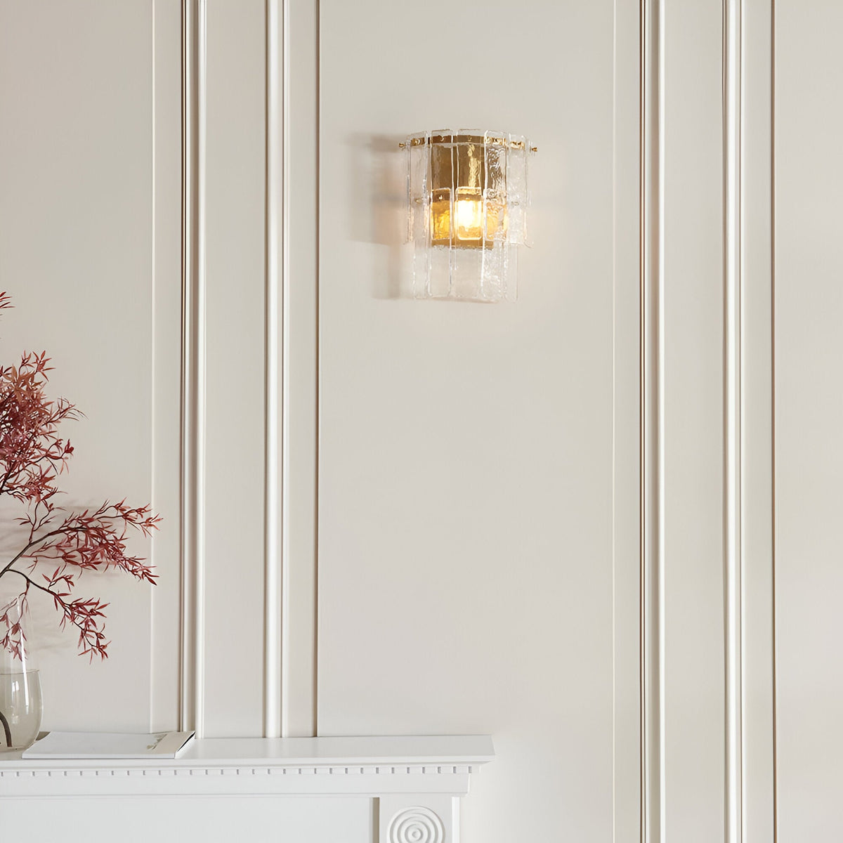 Brockhurst Wall Light in brass with layered textured glass panels, illuminated on a panelled wall above a mantelpiece with a vase of dried flowers nearby.
