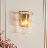 Brockhurst Wall Light in Brass, featuring layered textured glass panels on a brushed aged brass backplate, illuminated on a light wall beside foliage.
