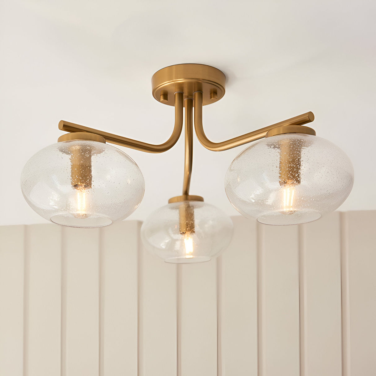 Pemberton 3 Light Semi-Flush Ceiling Light in brass with clear seeded glass globe shades, mounted on a panelled wall in a room setting.