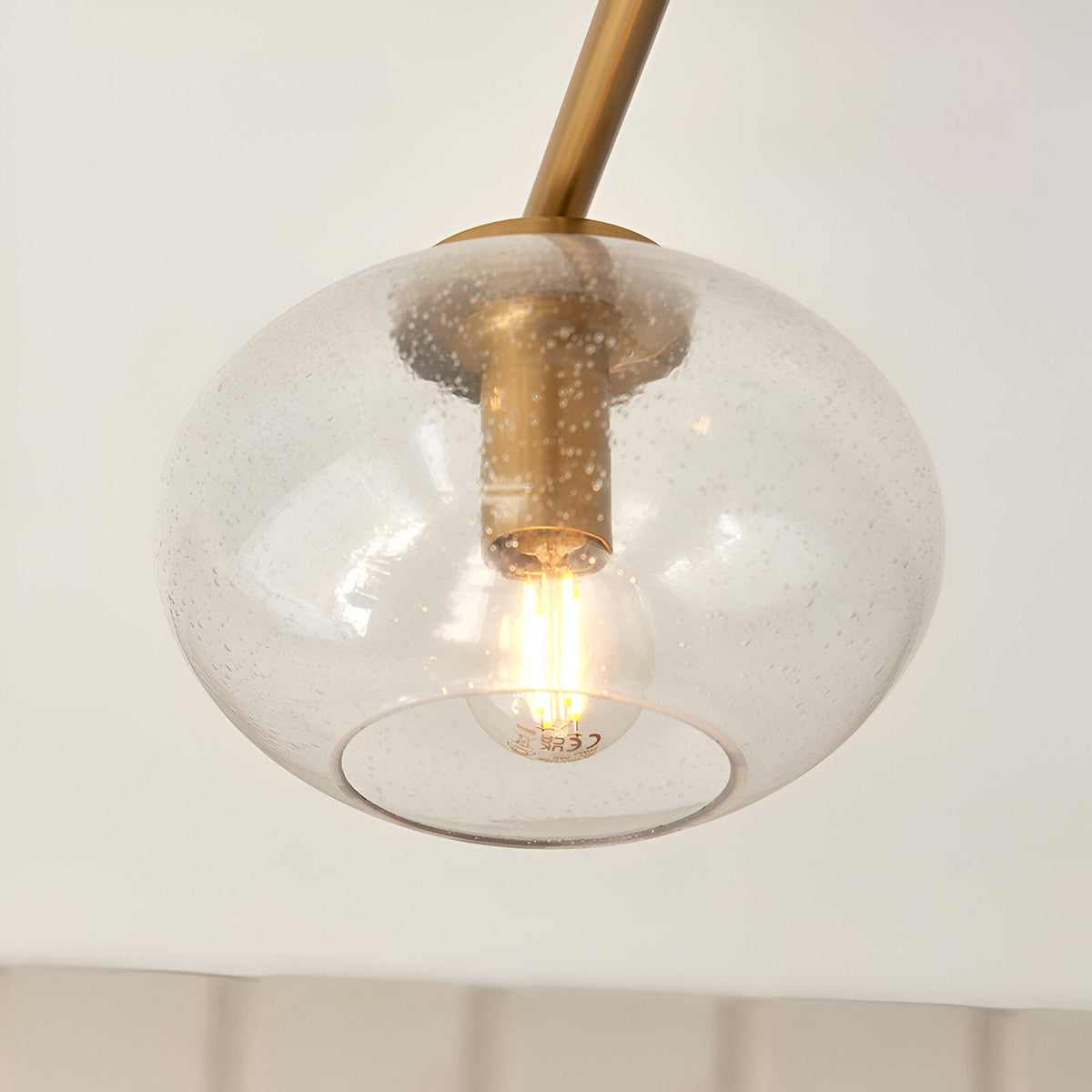 Pemberton 3 Light Semi-Flush Ceiling Light in brass, close-up of one seeded glass globe shade with lit bulb on a brass stem against a plain white background.