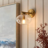 Endon Pemberton wall light in brass with a bubbled clear glass oval shade, mounted beside framed art on a panelled wall above a red-leafed plant.