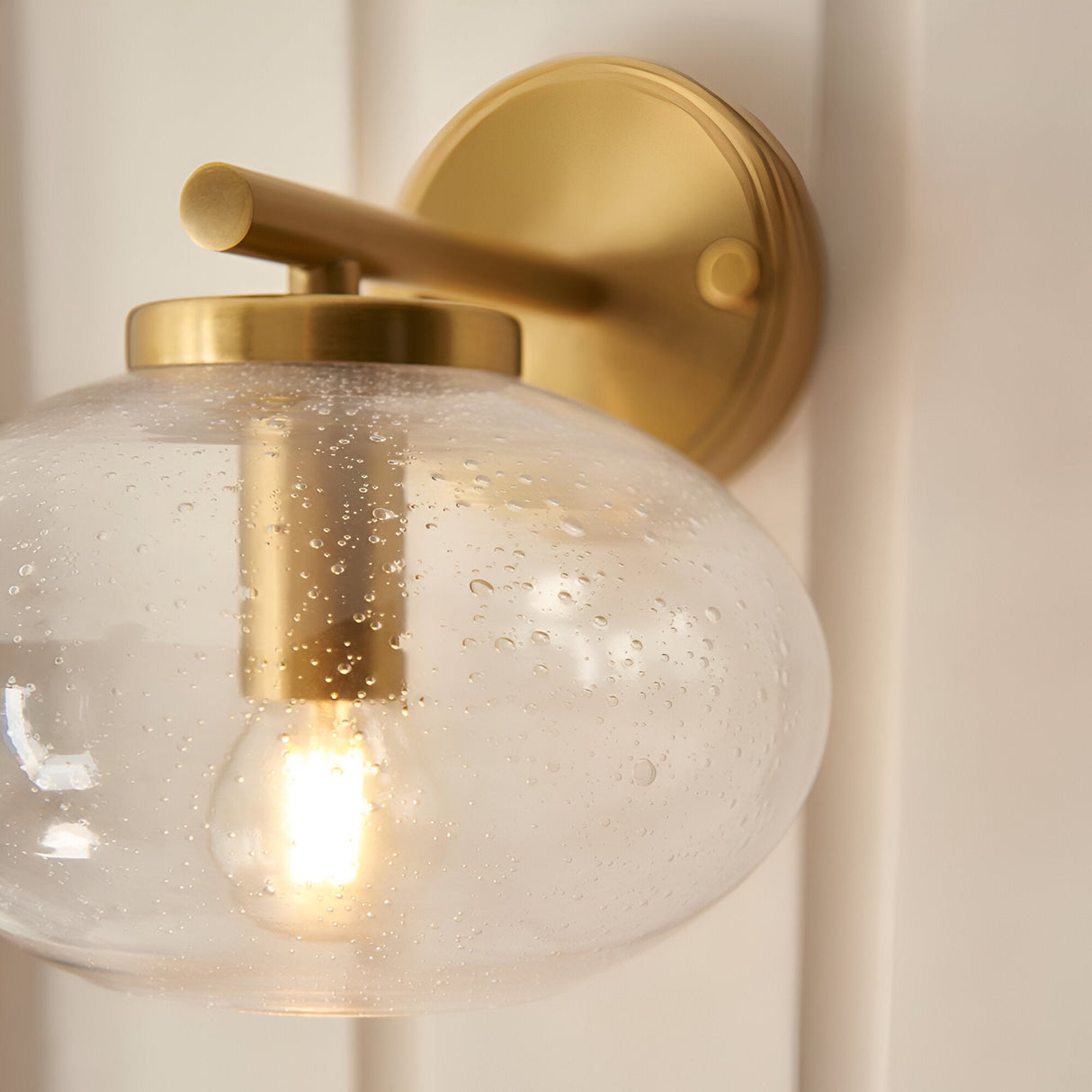 Close-up of the Endon Pemberton wall light showing the warm brass arm, backplate and hand-blown clear bubbled glass shade with lit E14 lamp.