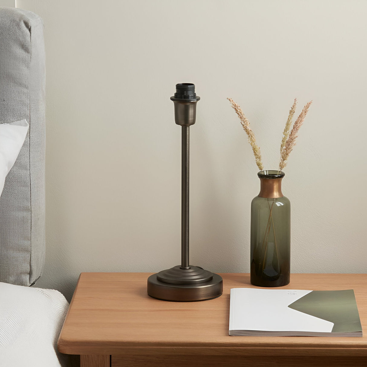 Endon Chartworth bronze table lamp base with stepped tapering stem and E14 fitting on a wooden bedside table beside a green glass vase with dried grasses.