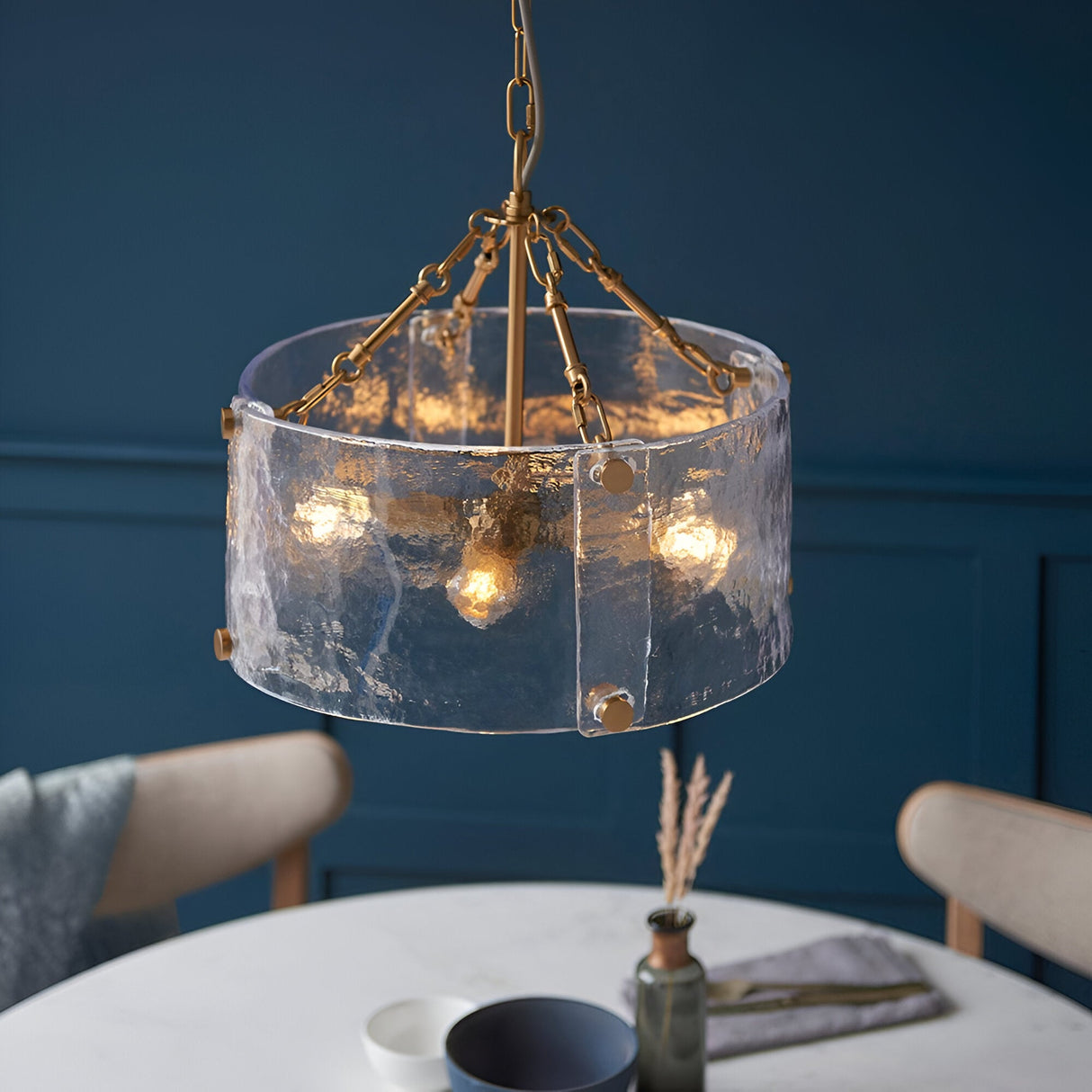 Brockmont 4 Light Pendant in brass with a cylindrical shade of textured glass panels, hung above a dining table in a dark blue-walled room.
