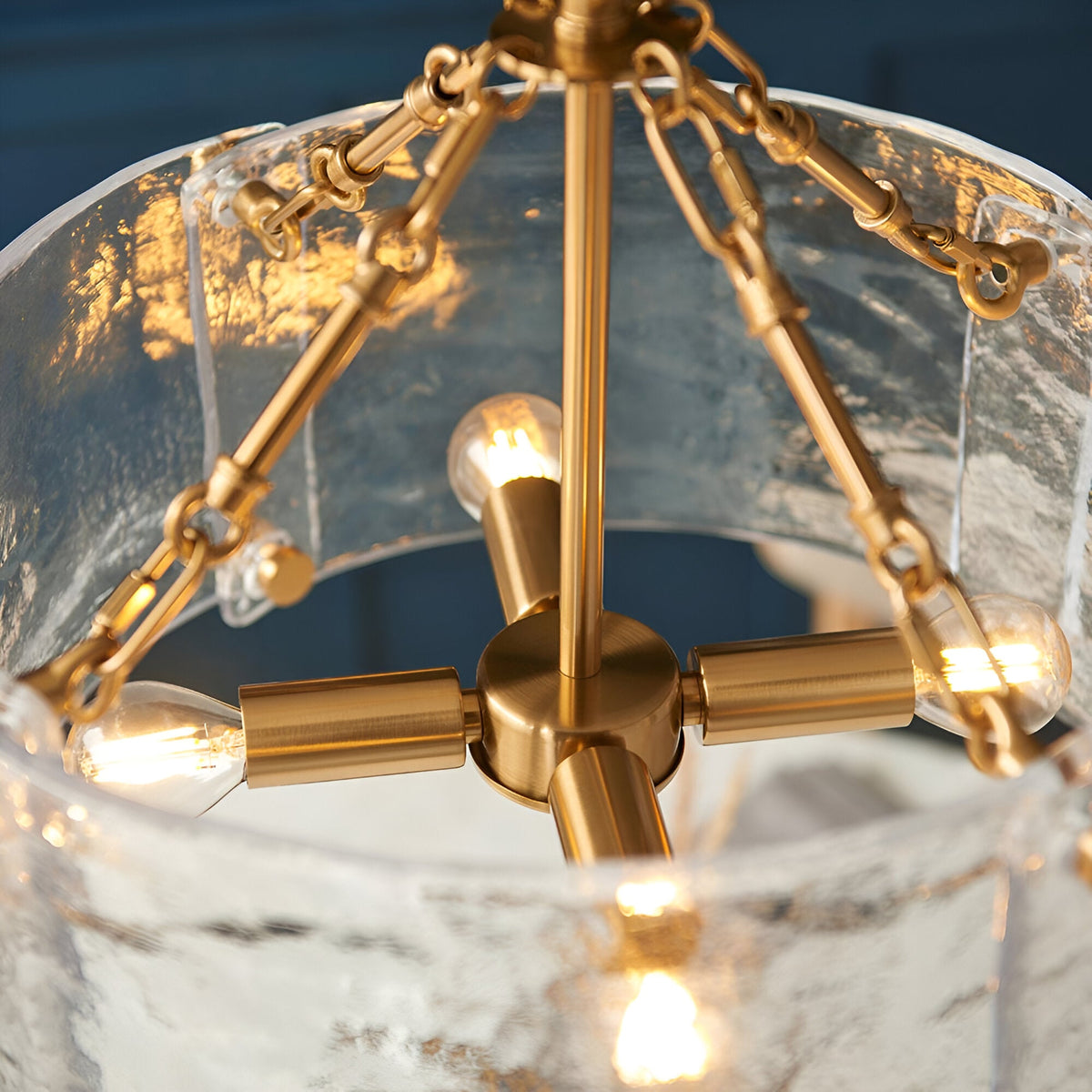 Close-up of the Brockmont 4 Light Pendant in brass, showing E14 bulbs, chain fittings, and textured glass shade panels.