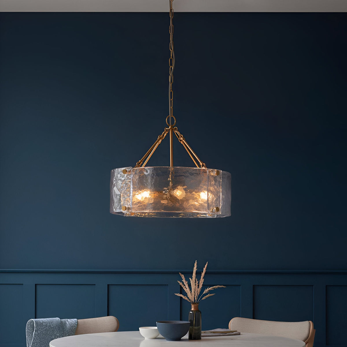 Endon Brockmont 6 light pendant with rippled handmade glass cylinder shade and brushed aged brass frame, hanging above a dining table against a deep blue wall.