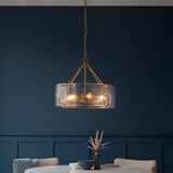 Endon Brockmont 6 light pendant with rippled handmade glass cylinder shade and brushed aged brass frame, hanging above a dining table against a deep blue wall.
