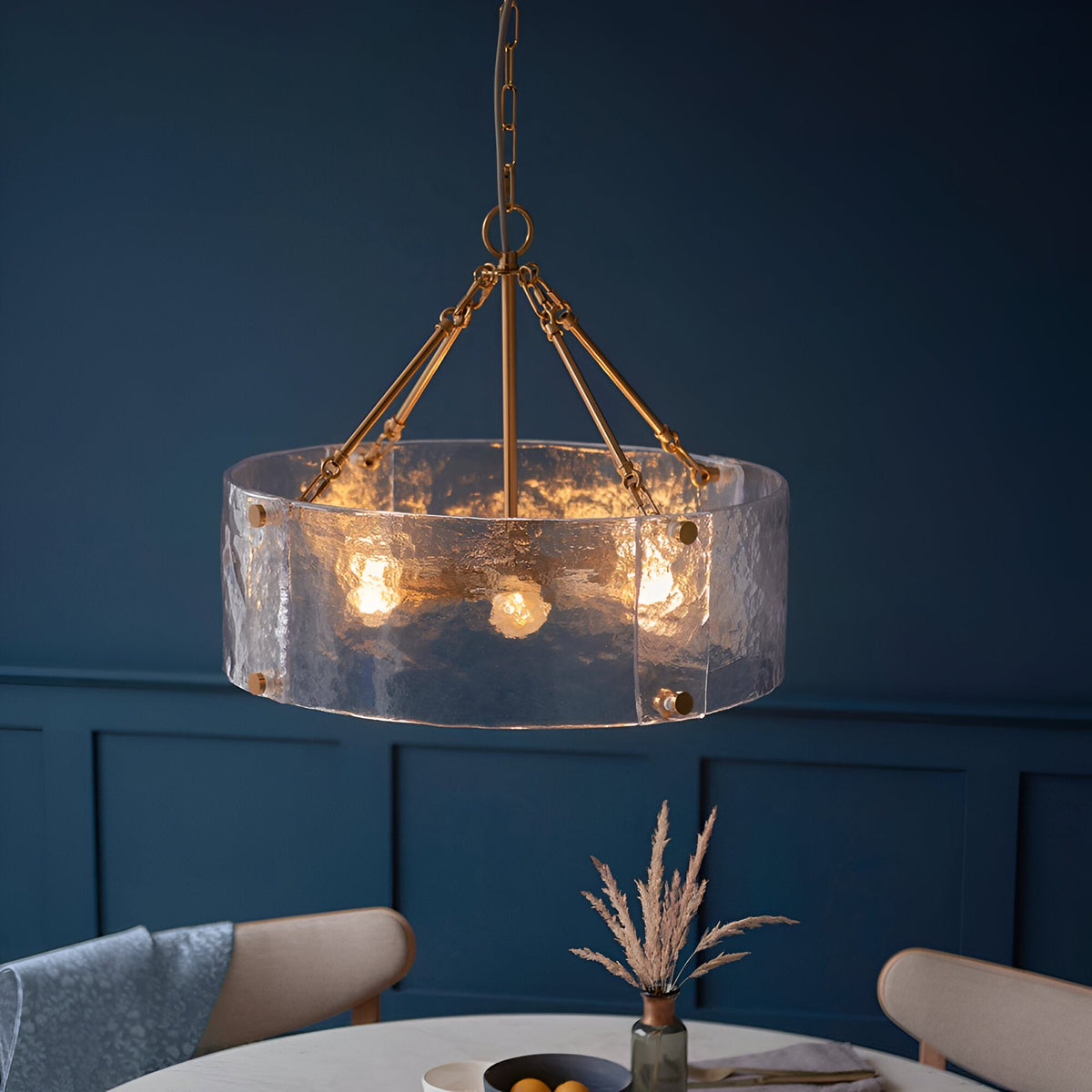 Endon Brockmont 6 light pendant with rippled handmade glass cylinder shade and brushed aged brass frame, hung above a dining table against a deep blue wall.