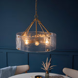 Endon Brockmont 6 light pendant with rippled handmade glass cylinder shade and brushed aged brass frame, hung above a dining table against a deep blue wall.