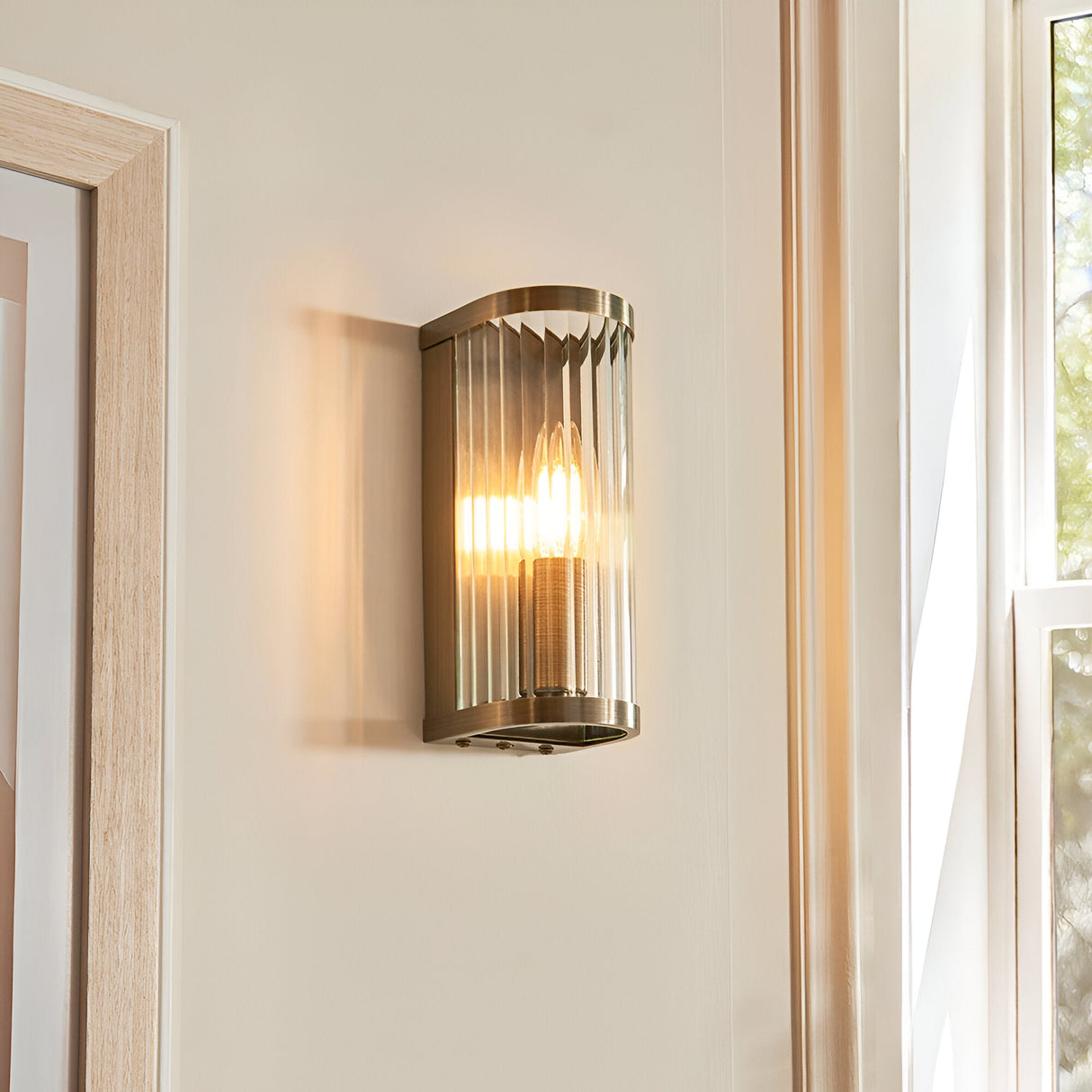 Endon Rothbury wall light in antique brass with vertically ribbed clear glass diffuser, illuminated on a hallway wall beside a doorway and window.