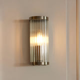 Endon Rothbury wall light in antique brass with a vertically ribbed clear glass diffuser, illuminated on a pale wall beside a panelled door frame.