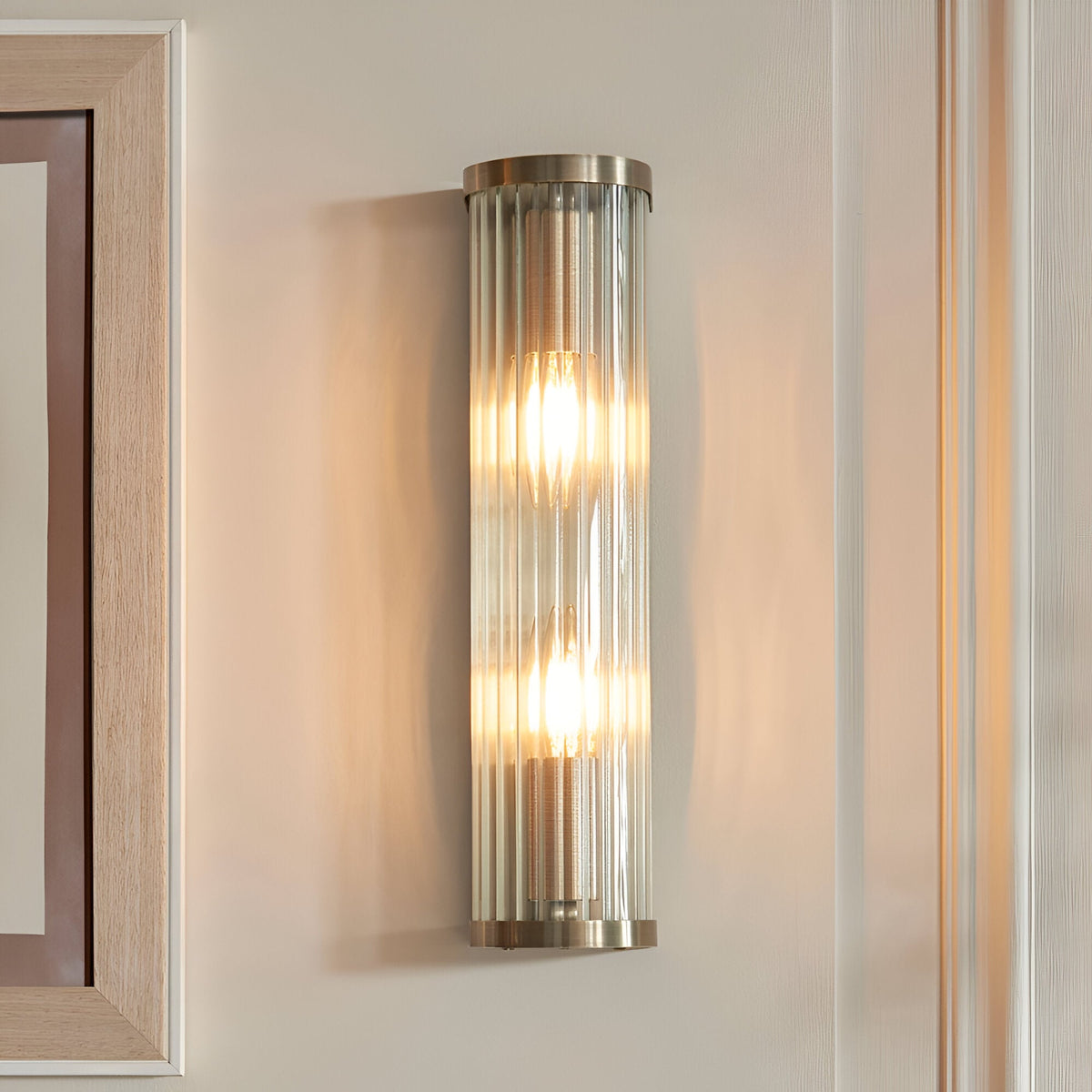Rothbury 2 Light Wall Light in antique brass with ribbed glass diffusers, mounted on a neutral wall, both bulbs illuminated.