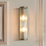 Rothbury 2 Light Wall Light in antique brass with ribbed glass diffusers, mounted on a neutral wall, both bulbs illuminated.