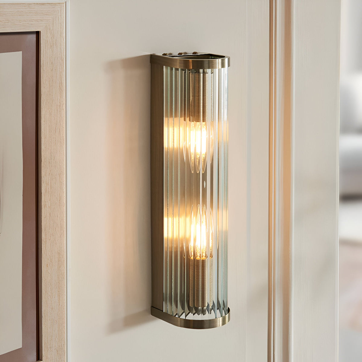 Rothbury 2 Light Wall Light in antique brass with vertically ribbed clear glass diffusers, shown illuminated on a wall beside a door frame.