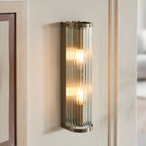 Rothbury 2 Light Wall Light in antique brass with vertically ribbed clear glass diffusers, shown illuminated on a wall beside a door frame.