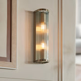 Rothbury 2 Light Wall Light in antique brass with ribbed glass diffusers and exposed bulbs, mounted on a cream panelled wall.