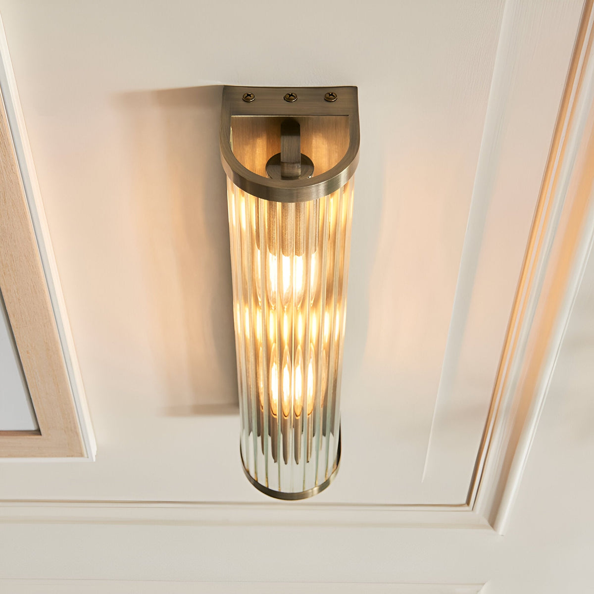 Rothbury 2 Light Wall Light in antique brass, featuring a ribbed cylindrical glass diffuser and curved bracket, mounted on a cream panelled wall.