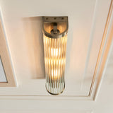 Rothbury 2 Light Wall Light in antique brass, featuring a ribbed cylindrical glass diffuser and curved bracket, mounted on a cream panelled wall.