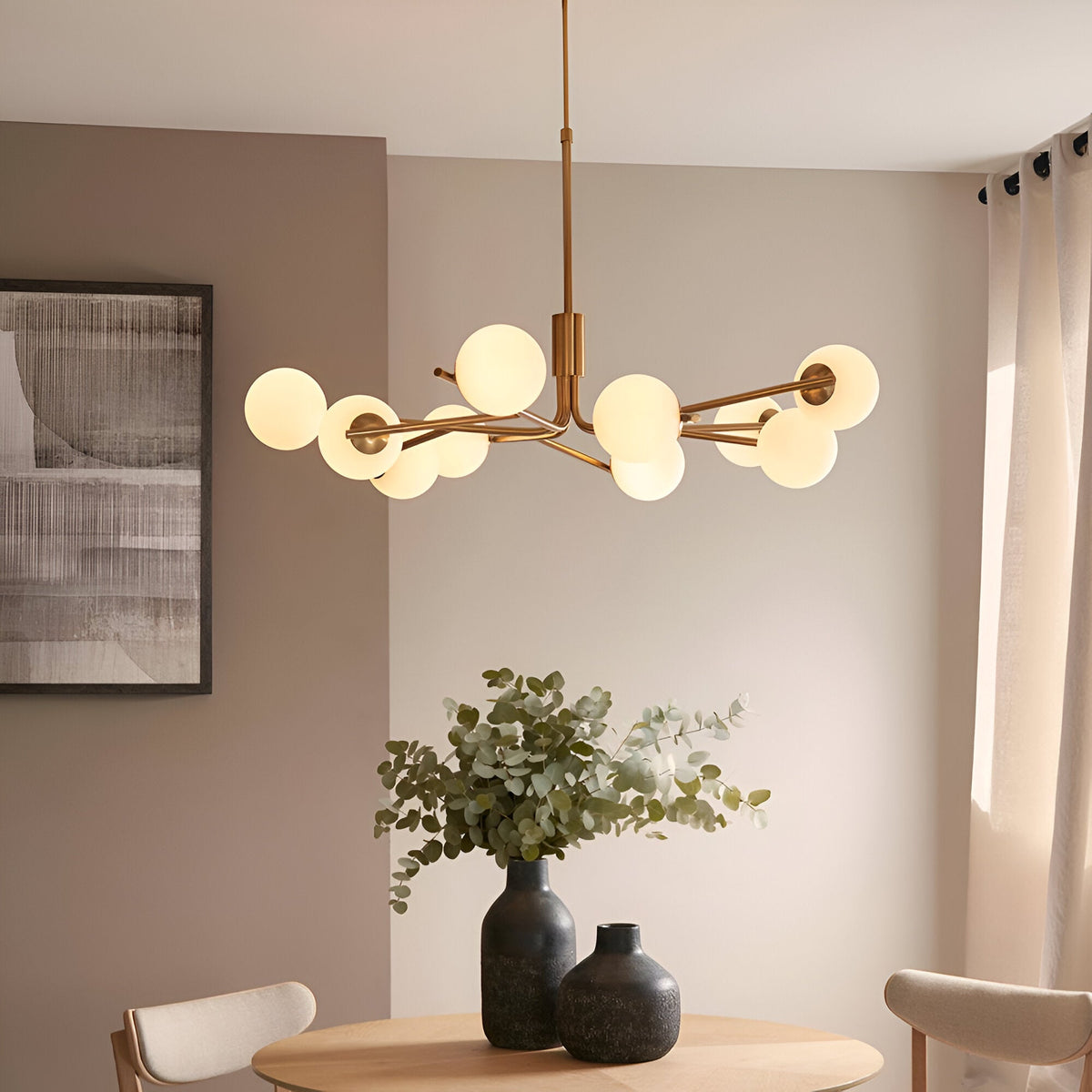 Grantham 10 Light Pendant in brushed aged brass with matt opal glass spheres on angled arms, shown above a dining table with vases and foliage.