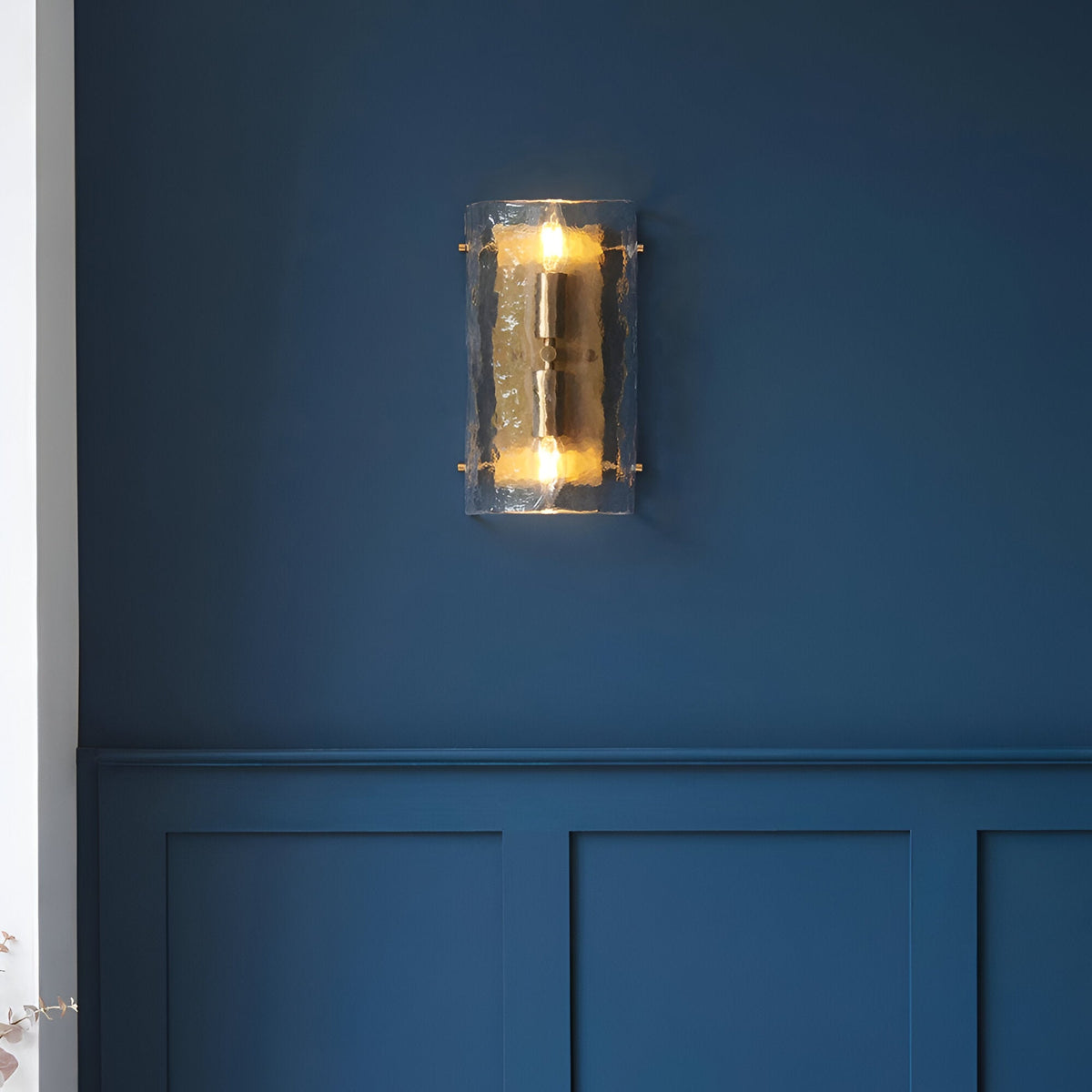 Endon Brockmont 2 light wall light illuminated on a deep blue panelled wall, with curved textured glass panel and brushed aged brass metalwork.