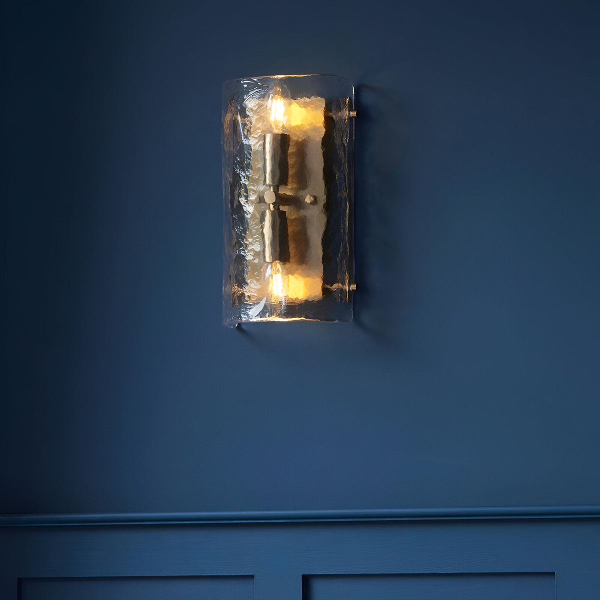 Endon Brockmont 2 light wall light with curved handmade textured glass panel and brushed aged brass metalwork, illuminated on a deep blue panelled wall.