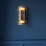 Endon Brockmont 2 light wall light with curved handmade textured glass panel and brushed aged brass metalwork, illuminated on a deep blue panelled wall.