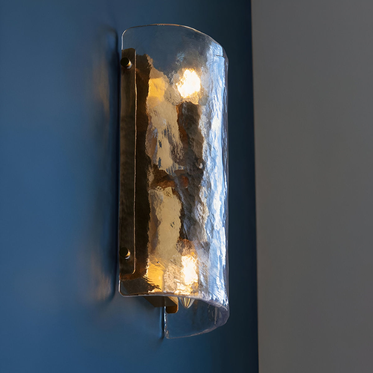Endon Brockmont 2 light wall light with curved handmade textured glass panel and brushed aged brass metalwork, illuminated on a dark blue wall.