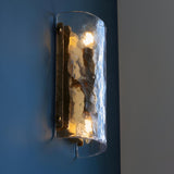 Endon Brockmont 2 light wall light with curved handmade textured glass panel and brushed aged brass metalwork, illuminated on a dark blue wall.