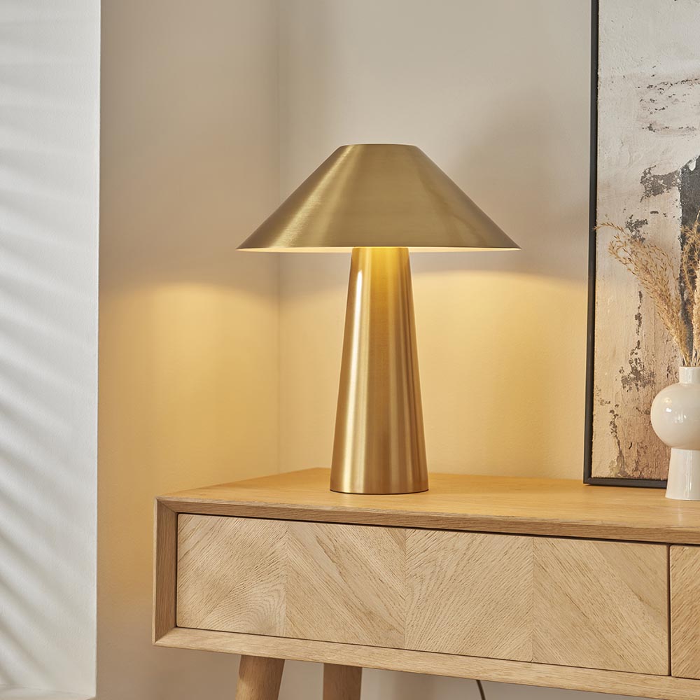 Endon Brockbourne table lamp in brushed antique brass with a wide conical shade and tapered base, styled on a light oak sideboard beside framed art.