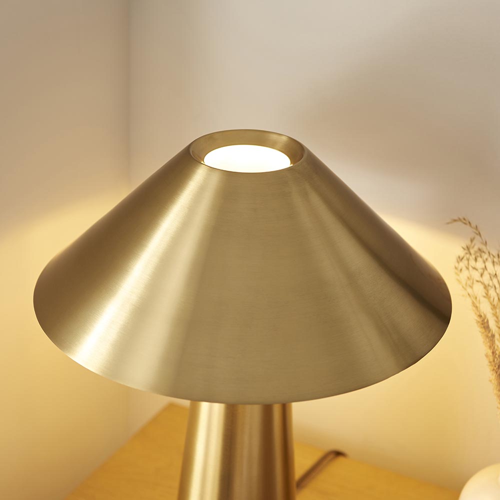 Endon Brockbourne table lamp with a wide, low brushed antique brass shade and cylindrical base, lit on a wooden surface beside dried grasses.