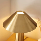 Endon Brockbourne table lamp with a wide, low brushed antique brass shade and cylindrical base, lit on a wooden surface beside dried grasses.